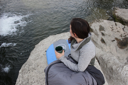 Woman enjoying winter camping hygiene tips while relaxing by the water, wrapped in a sleeping bag. using roman trail outfitters