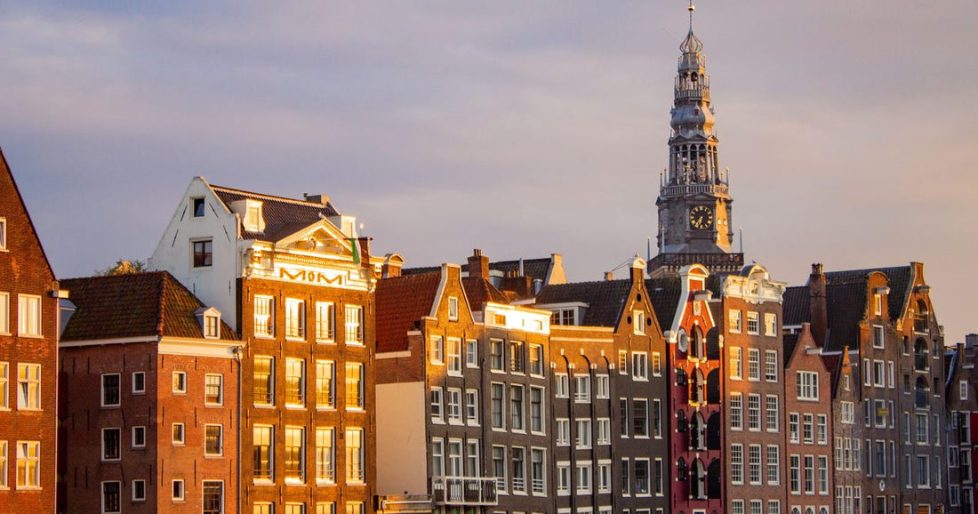 Classic Amsterdam gabled canal houses glowing warm gold at sunset with a church spire, women's Amsterdam packing list by Roman Trail Outfitters