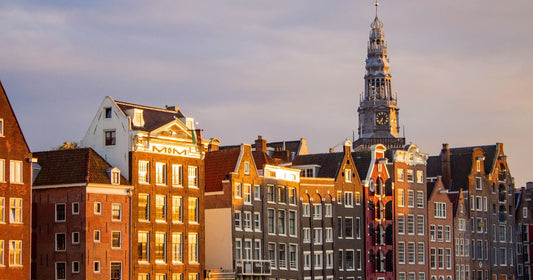 Classic Amsterdam gabled canal houses glowing warm gold at sunset with a church spire, women's Amsterdam packing list by Roman Trail Outfitters