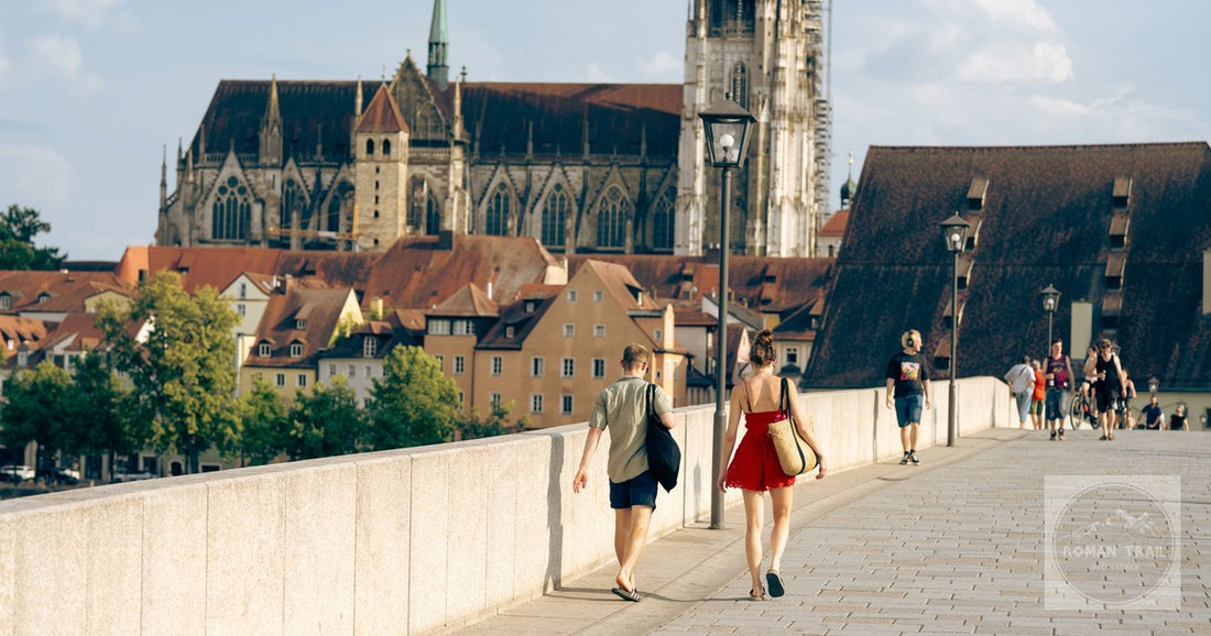 People walking on Steinerne Brücke with Regensburg Cathedral in the background on a sunny day.