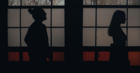 Silhouettes of a man and woman standing apart, divided by a window frame, facing opposite directions
