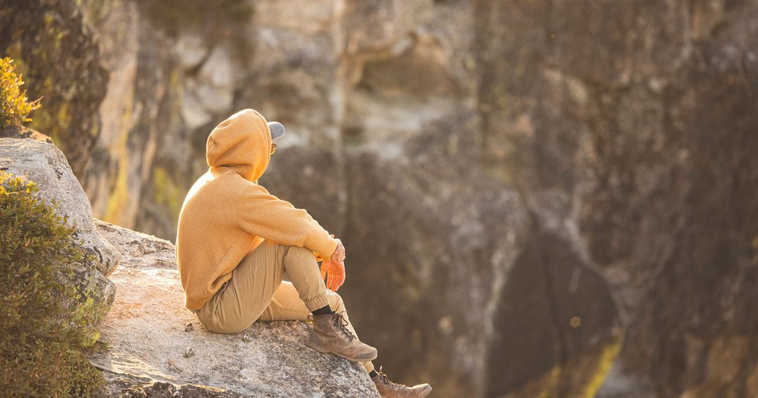 A man sitting still on a cliff edge in warm golden light, hands resting on his knees, overlooking a canyon