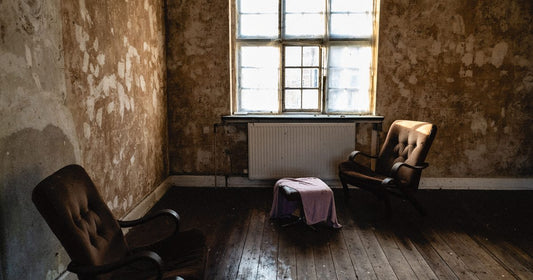 Two empty weathered chairs facing a sunlit window in a worn room, a decision not yet made