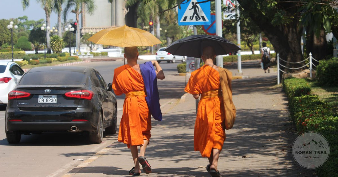 Two Buddhist monks walking a sunny sidewalk in Vientiane, Laos, holding umbrellas for shade