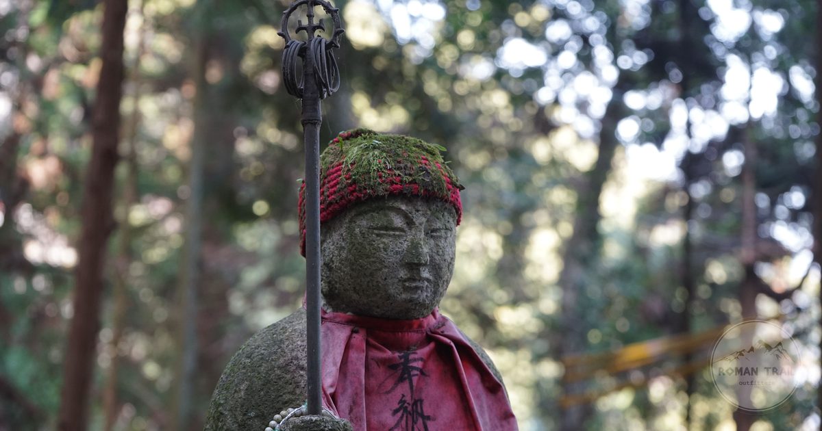 A moss covered stone Jizo statue in a red cloth in a forest cemetery, Japan