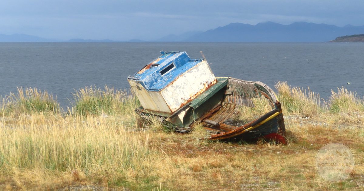 A worn boat on a grassy shore with the sea and mountains at Punta Arenas, Chile