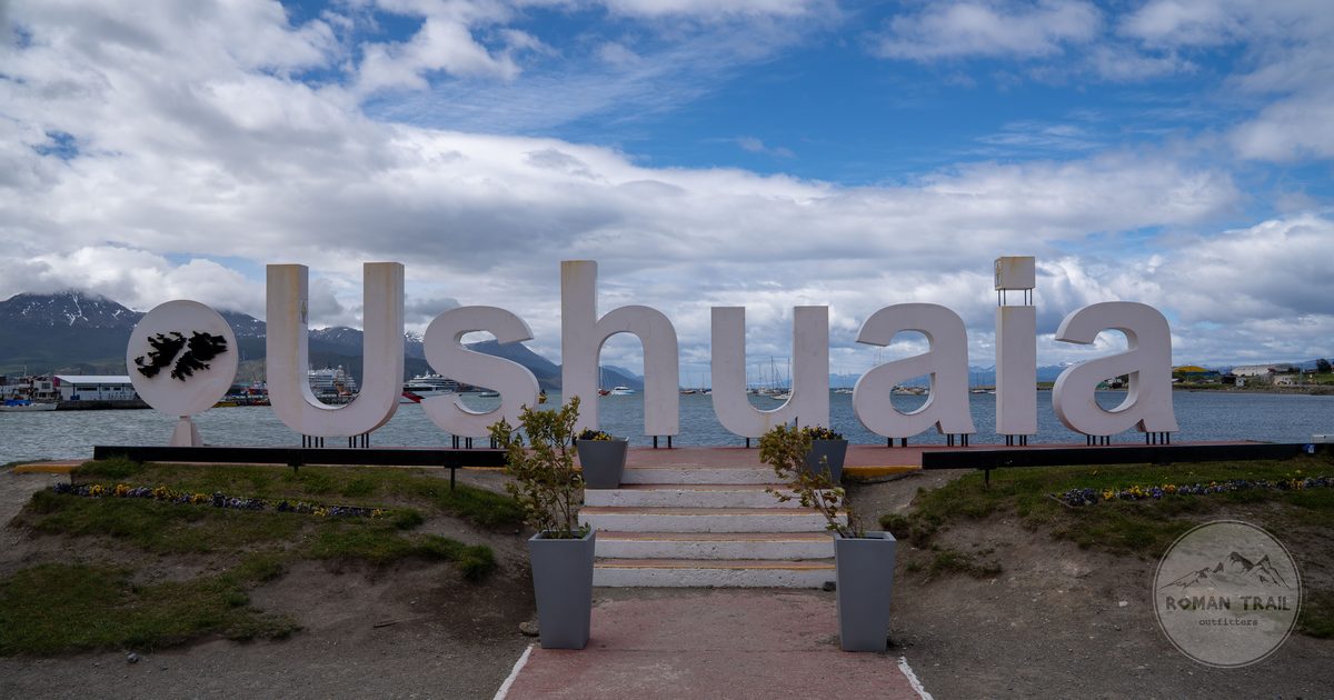 The Ushuaia end of the world sign with the Beagle Channel and mountains behind it, Ushuaia packing list for women by Roman Trail Outfitters