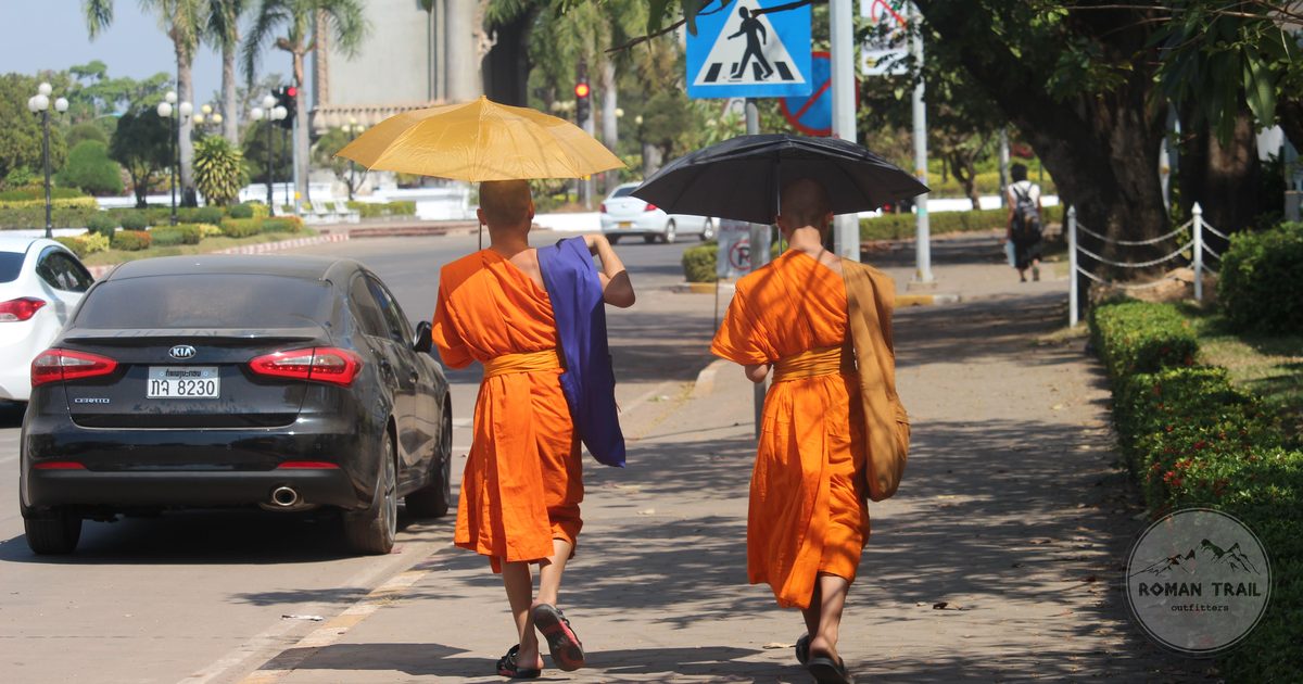 Two Buddhist monks walking a sunny sidewalk in Vientiane, Laos, holding umbrellas for shade